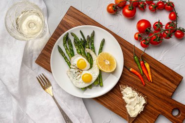 Mutfak masasında, ahşap mutfak tahtasında iki omlet, kızarmış kuşkonmaz ve zeytin ile birlikte beyaz bir tabak var. Yakında taze vişneli domates, acı biber ve yumuşak peynirli tost var. Beyaz şaraplı bardak..