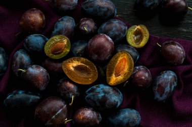 Koyu olgun sulu erikler mor bir masa örtüsünün üzerinde uzanır, siyah ahşap arka planda yatar. Birkaç tekne kesilmiş..