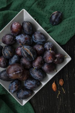 Koyu olgun sulu erikler, yeşil bir masa örtüsünün üzerindeki gri kare bir tabakta uzanır. Siyah ahşap arka planda yatar. Tabağın yanında birkaç kemik var..