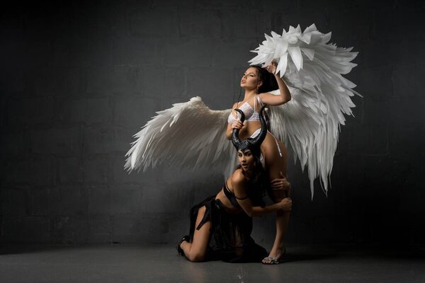 Graceful woman in underwear outstretching feather wings while putting leg on sexy female model wearing devil fairy costume in studio