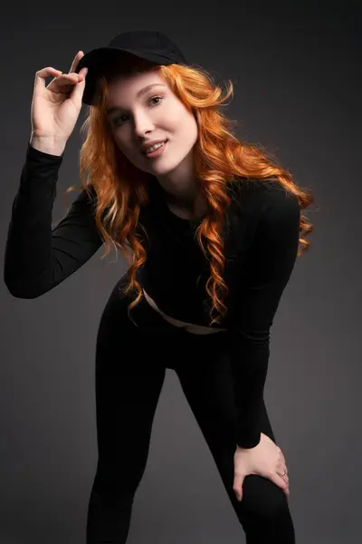 Self assured young female model with curly long red hair in trendy activewear adjusting cap and looking at camera while leaning forward against gray background