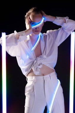 Makyajlı beyaz gömlekli, pantolonlu, kendine güvenen genç çift cinsiyetli erkek manken yüzüne dokunuyor ve LED çubukların yanındaki siyah stüdyoda dikilirken kameraya bakıyor.