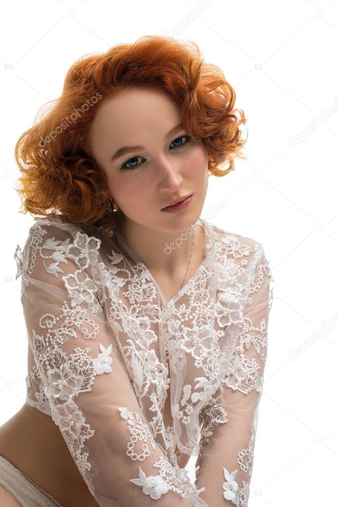 Gentle female model with curly red hair wearing transparent lace cropped top on bare breast and underpants looking at camera against white background