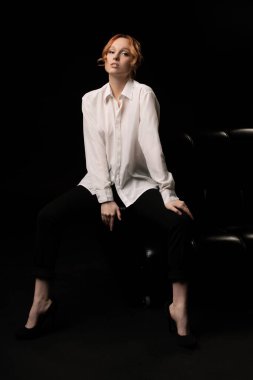 Kendine güveni tam, kızıl saçlı, şeffaf beyaz gömlekli, rahat deri koltukta oturan, kendine güvenen genç bir bayan menajer.