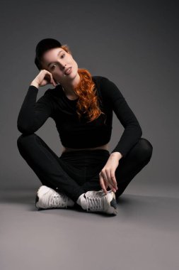 Uzun kızıl saçlı, şık spor elbiseli ve spor ayakkabılı, yerde oturmuş kameraya bakan rahat ve genç bir kadın vücudu.