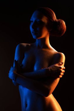 Serin ve sıcak ışıkların altında üstsüz bir kadın ıslak cilde su damlatarak göğsüne dokunuyor.