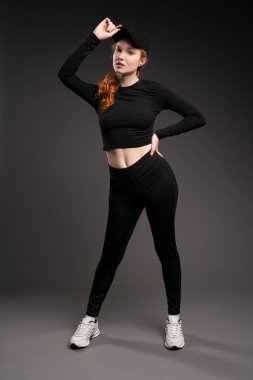 Kendine güvenen, kıvırcık, uzun kızıl saçlı, modaya uygun bir şapka takan ve gri arka plana yaslanarak kameraya bakan genç bir model.