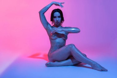 Uzun siyah saçlı, parlak vücutlu, aydınlık stüdyoda oturan seksi bir kadın.