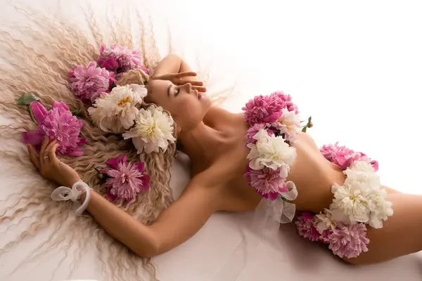 High angle of alluring young fit female with fresh flowers on naked body and long blond curly hair lying on comfortable white bed in daylight