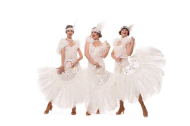 Beyaz sanat deco kostümlü, saç bandı takmış, beyaz arka planda tüy yelpazeli kameralara bakan içerik sahibi kadın dansçılar.