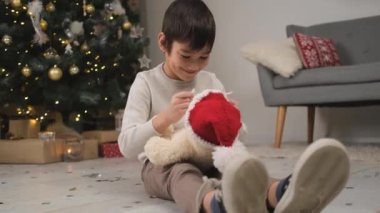 Küçük çocuk Noel kutlaması için dekore edilmiş odada oyuncak ayıya örülmüş Noel Baba şapkası takıyor.