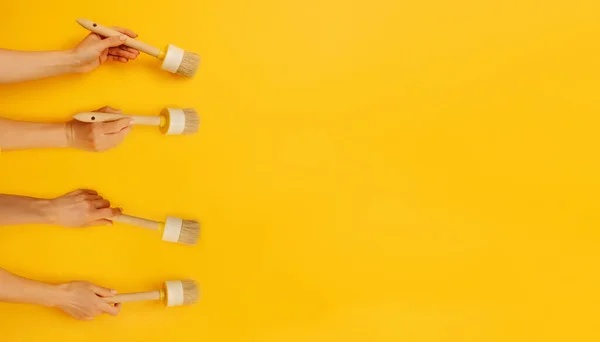 Four hands holding paint brushes on yellow background with copy space. Concept of creativity, improvement and inspiration. Working tool equipment for brushing