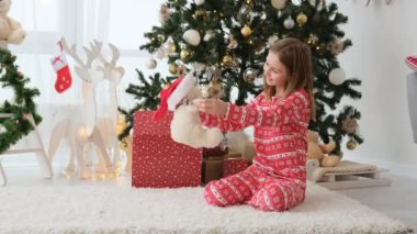 Pretty child girl playing with gift teddy bear next to Christmas tree. Beautiful female kid enjoying present toy in New Year and Xmas time