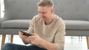 Adam yerde oturuyor, akıllı telefonda oynuyor ve gülümsüyor. Yetişkin adam cep telefonuna bakıyor ve mobil oyunlarla eğleniyor.