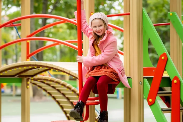 秋の屋外で遊び場に座っているかわいい女の子の子供とカメラを見ています 公園で放課後を楽しむ女性の子供 — ストック写真