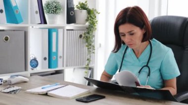 Klinikte oturan, not defterine not alan ve elinde siyah dosya tutan kadın doktor portresi. Coronavirus zamanındaki hastanede sağlık uzmanı. Mavi üniformalı doktor.
