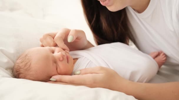 漂亮的年轻母亲穿着白色布衣 带着新生婴儿的特写 漂亮的女孩在床上让睡着的婴儿平静下来 微笑着 小孩打瞌睡和他妈妈关心小孩 — 图库视频影像
