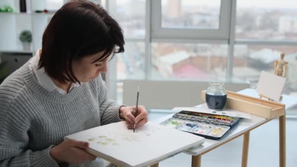 女人坐在桌旁 用水彩画着树叶 用彩绘工具在光线室工作室里的白纸上 女性用水彩画来创作艺术作品 — 图库视频影像