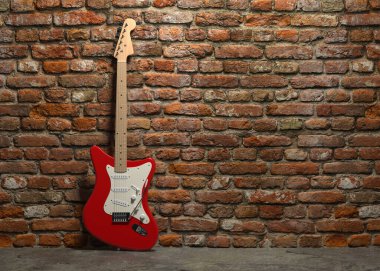 Altı telli, tahta gövdeli, kırmızı elektro gitar tuğla bir duvarın arkasında duruyor..