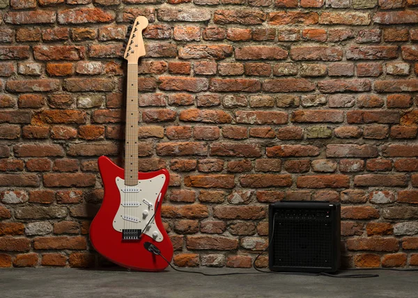 Red electric guitar with six string wooden body and fretboard wired to combo amp stands against the background of a brick wall.