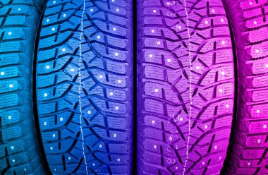 Winter studded tire in blue and pink tones. Winter car tires background. Tire stack background. Tyre protector close up. Square powerful spikes. Black studdable winter tyre profile. Car tires 