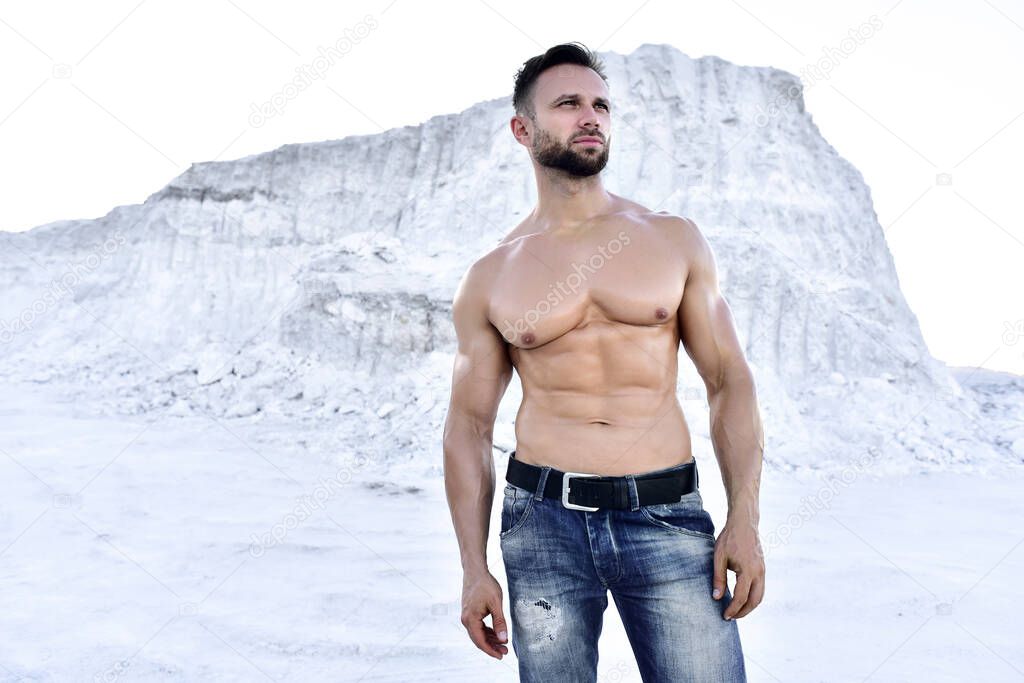 athletic man with a naked torso and jeans stands against the backdrop of a mountain