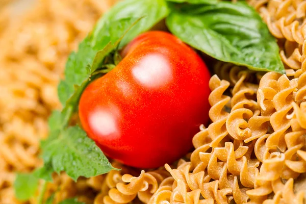 Einzelne reife Tomaten in einem Haufen ungekochter Nudeln und Basilikum — Stockfoto