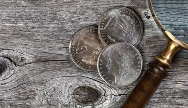 Vintage collection of US Morgan silver dollars with magnifying glass on rustic wooden boards in flat lay view clipart