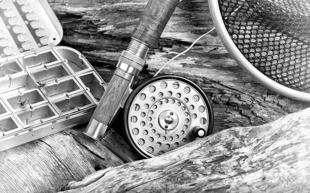 Antique fly rod, reel, landing net and lure container in stone and drift wood riverbed background with vintage effect.