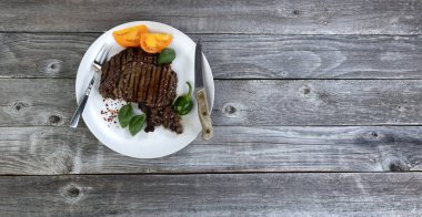 Düz bir ahşap masanın üzerinde otlar ve sebzelerle taze ızgara biftek. 