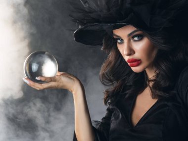 Woman in Halloween witch costume future teller showing crystal ball. Beautiful sexy model girl in sinister costume and make up over foggy background clipart