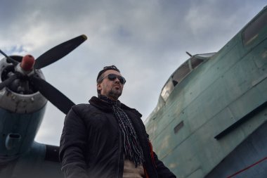 Bombacı ceketli eski okul pilotu. Uçağın yanında poz veriyor. Atkı ve güneş gözlüklü havacı erkek.
