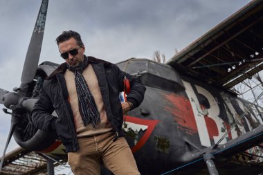 Bombacı ceketli eski okul pilotu. Uçağın yanında poz veriyor. Atkı ve güneş gözlüklü havacı erkek.