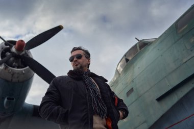 Bombacı ceketli eski okul pilotu. Uçağın yanında poz veriyor. Atkı ve güneş gözlüklü havacı erkek.