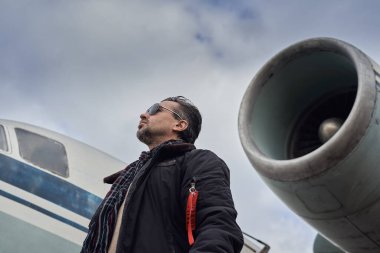 Bombacı ceketli eski okul pilotu. Uçağın yanında poz veriyor. Atkı ve güneş gözlüklü havacı erkek.