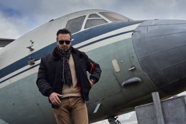 Bombacı ceketli eski okul pilotu. Uçağın yanında poz veriyor. Atkı ve güneş gözlüklü havacı erkek.