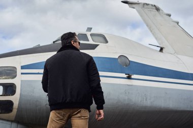 Bombacı ceketli eski okul pilotu. Uçağın yanında poz veriyor. Atkı ve güneş gözlüklü havacı erkek.