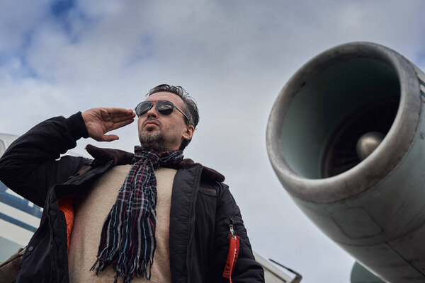 Oldschool pilot in the bomber jacket posing near the aircraft, aviator male with scarf and sunglasses