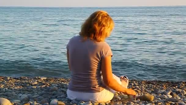 Sahilde deniz kenarında oturan ve suya taş atan bir kadın — Stok video