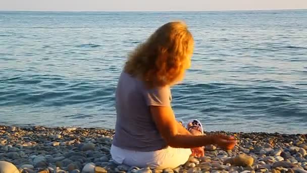 Een vrouw zittend op het strand door de zee en het gooien van stenen in het water — Stockvideo