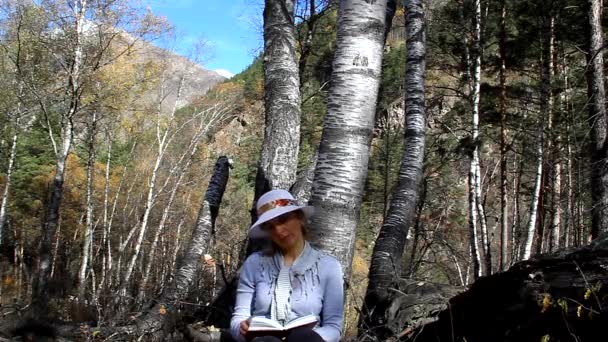 Femme au chapeau assise sous un arbre et lisant un livre — Video