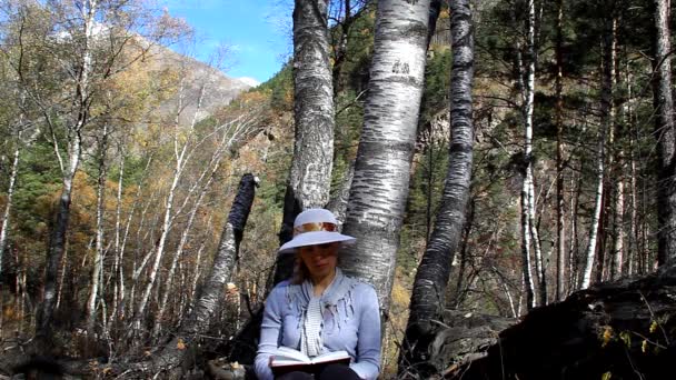 Женщина в шляпе сидит под деревом и читает книгу — стоковое видео