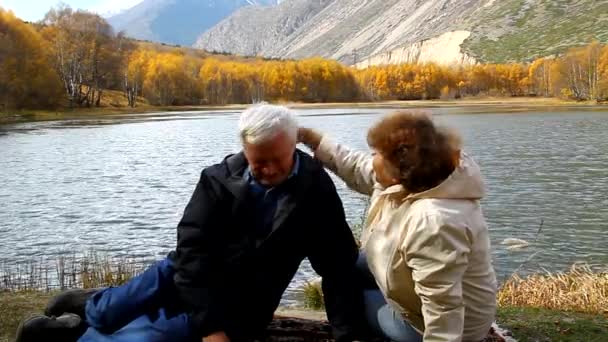 Happy mature man and woman sitting on the shore of the lake and pat each other on the head — Stock Video