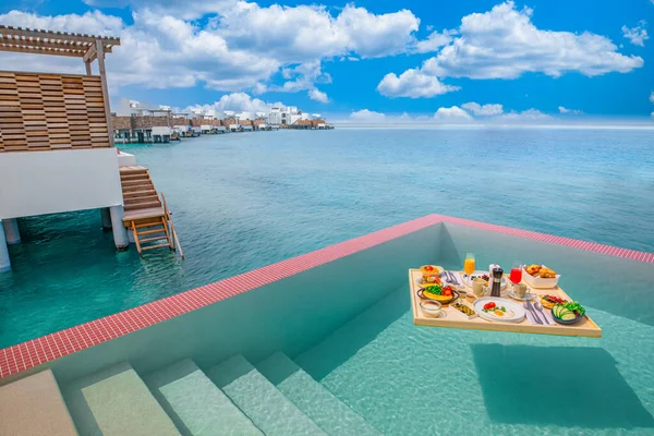 Breakfast in swimming pool, floating breakfast in luxurious tropical resort. Table relaxing on calm pool water, healthy breakfast and fruit plate by resort pool. Tropical couple beach luxury lifestyle