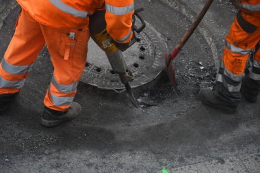 Koruyucu giysili işçiler rögar kapağında yol inşaatında pnömatik çekiçle çalışıyorlar.