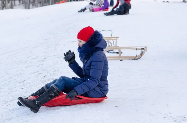 Girl sit on sled with blue jacket, camera in hand — Stock Photo, Image