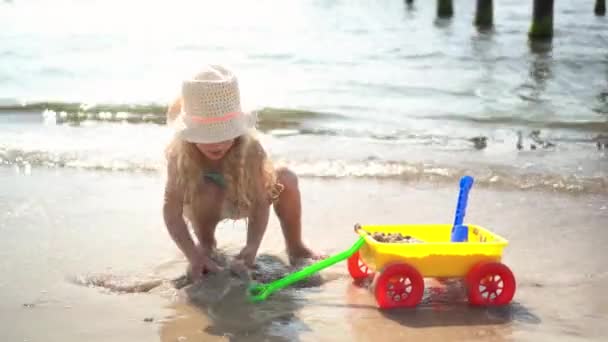 Cute girl z kręconymi blond włosy bawiąc się z piasek mrówka zabawka koszyk na brzegu morza — Wideo stockowe
