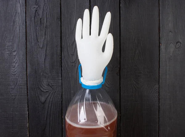 The production of wine. A plastic bottle with a young homemade wine and put on the neck of a rubber glove for fermentation. On a black wooden background. — Stock Photo, Image