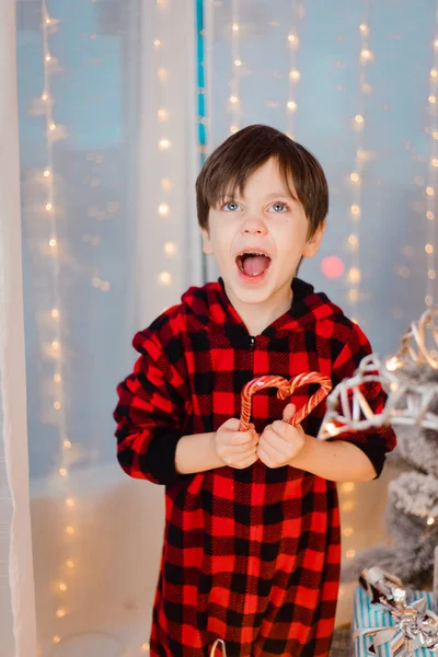 Chlapec v pyžamu s karamelem pod vánočním stromečkem. Novoroční bonbóny jsou červené. — Stock fotografie