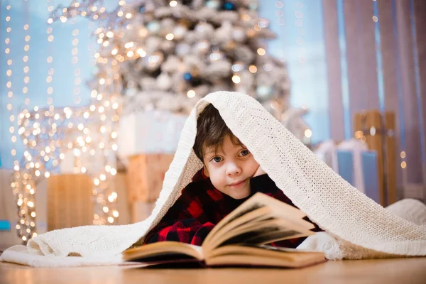Seorang anak laki-laki dengan piyama membaca buku di bawah pohon Natal. Suasana tahun baru. Membaca buku. Buku Childrens. Stok Gambar